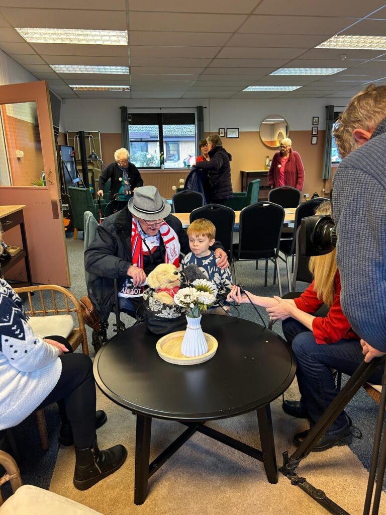 Eddie, wearing a Liverpool scarf, sitting with his grandson Harry and Shankley the toy dog while an ITV camera films them